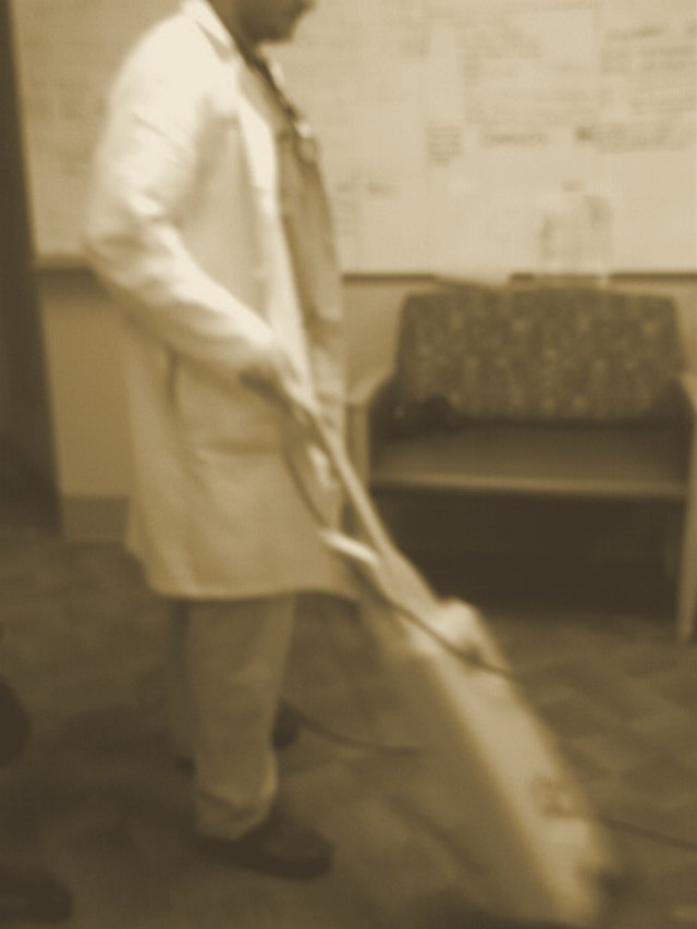 Rory as a resident in a white coat over scrubs, cleaning the workroom floor