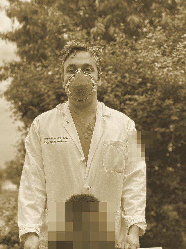 Rory in a mask and white coat standing behind his young child, whose face is blurred for privacy